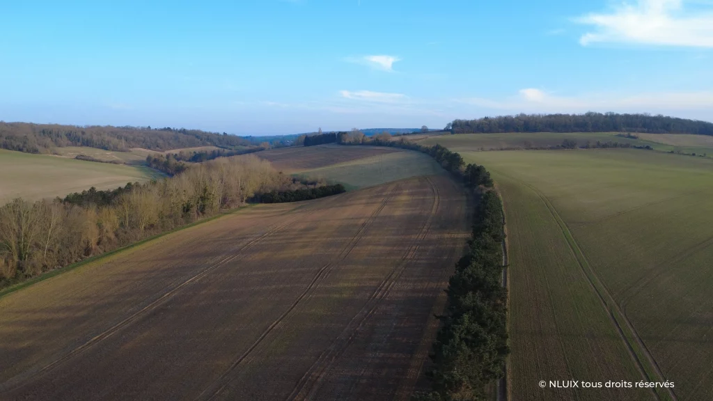 Capture aérienne du paysage dans le Vexin. La séparation visuelle et courbée vient séparer des parcelles agricoles et guide le regard vers l'horizon bleu légèrement vallonné. 